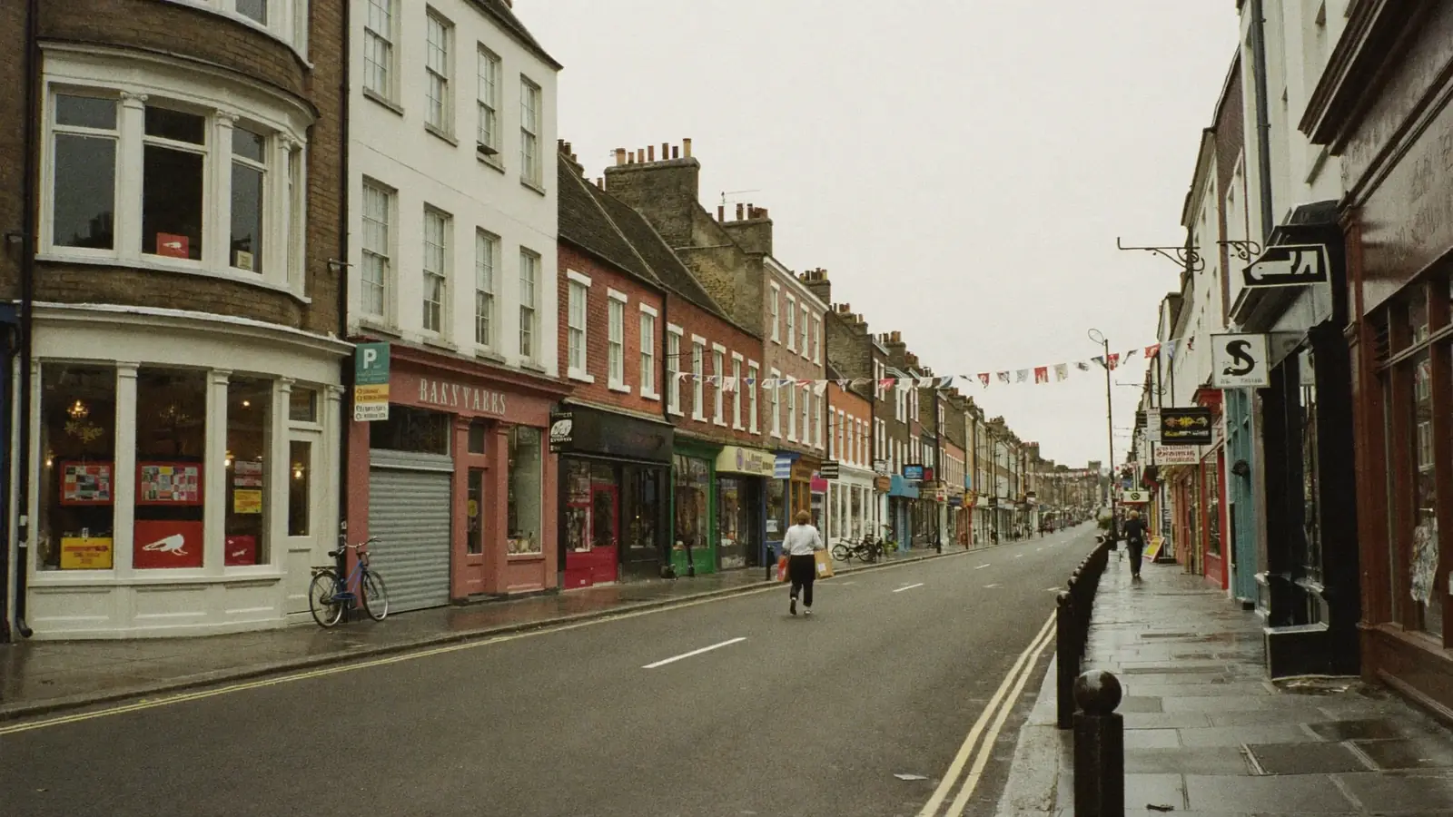 A British high street on an overcast day, with shops, a cyclist's bike by the kerb and a shopper walking away